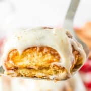 A close-up of a frosted cinnamon roll on a spatula, showing layers of dough and icing reminiscent of soft Hawaiian Roll texture, with more cinnamon rolls blurred in the background.