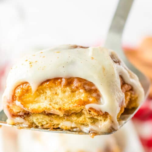 A close-up of a frosted cinnamon roll on a spatula, showing layers of dough and icing reminiscent of soft Hawaiian Roll texture, with more cinnamon rolls blurred in the background.