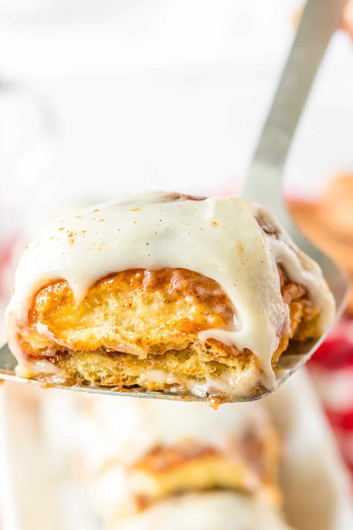A close-up of a frosted cinnamon roll on a spatula, showing layers of dough and icing reminiscent of soft Hawaiian Roll texture, with more cinnamon rolls blurred in the background.