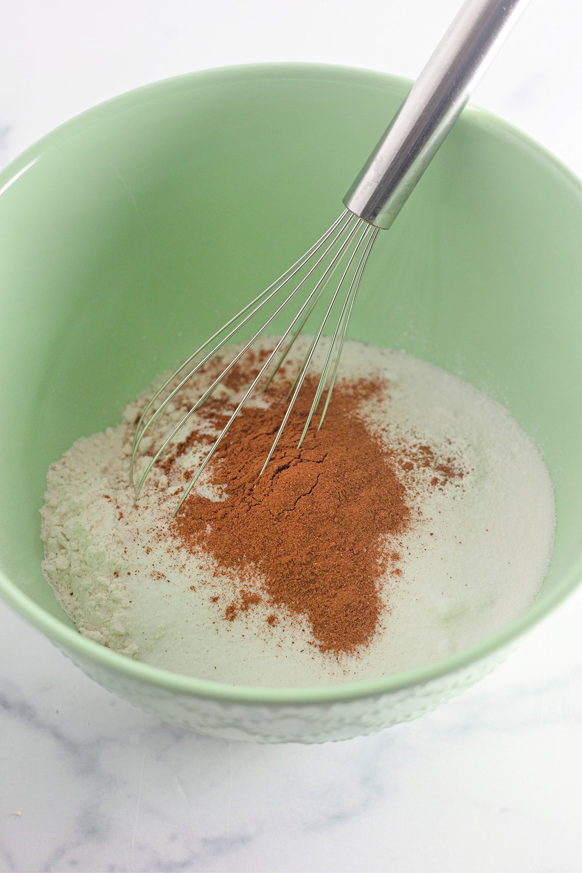 Green bowl containing flour, sugar, and cinnamon being mixed with a metal whisk on a white surface—perfect as the base for a delicious Pumpkin Wacky Cake.