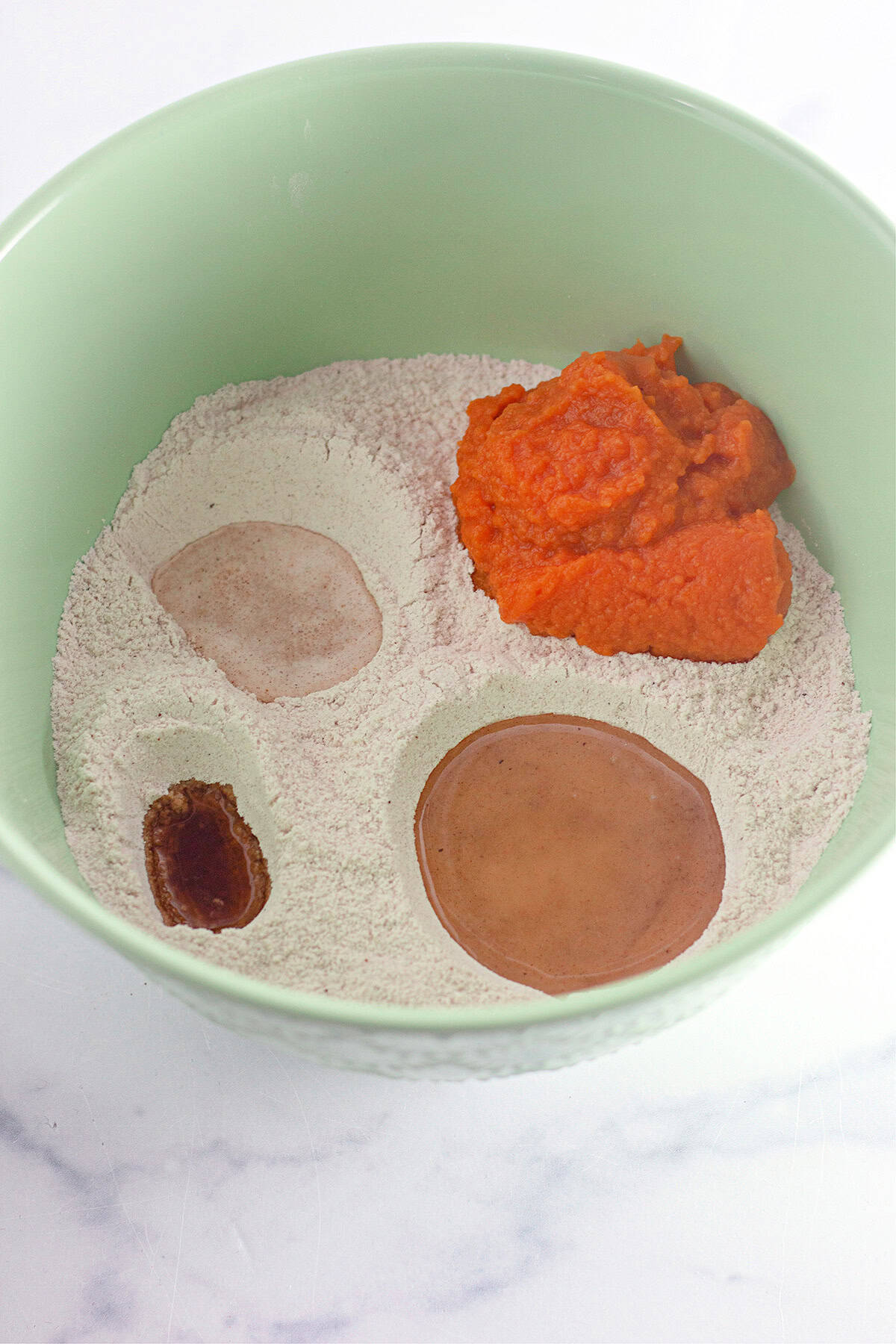 Green bowl containing flour, brown liquid, pumpkin puree, and another brown substance, all unmixed, on a white marble surface—the perfect beginning for a delicious Pumpkin Wacky Cake.