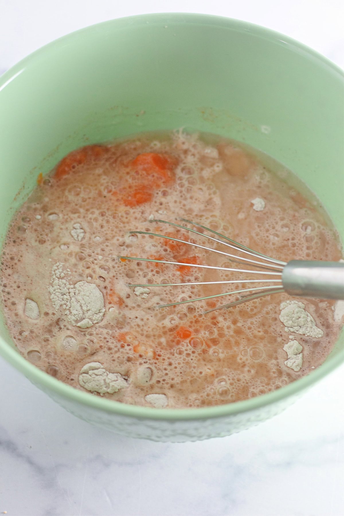 A light green bowl with a metal whisk mixing flour, orange liquid, and water for Pumpkin Wacky Cake. The mixture appears partially combined with bubbles on the surface, capturing the early steps of this unique cake recipe.