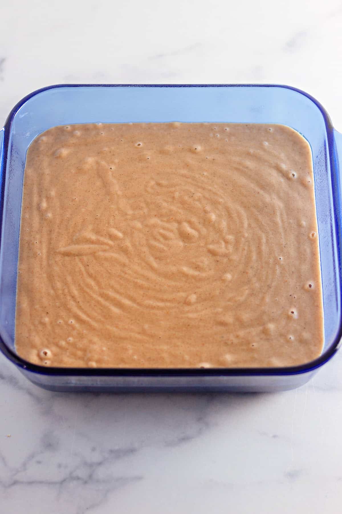 A square glass container filled with a light brown, smooth Pumpkin Wacky Cake batter sits invitingly on a white marble surface.