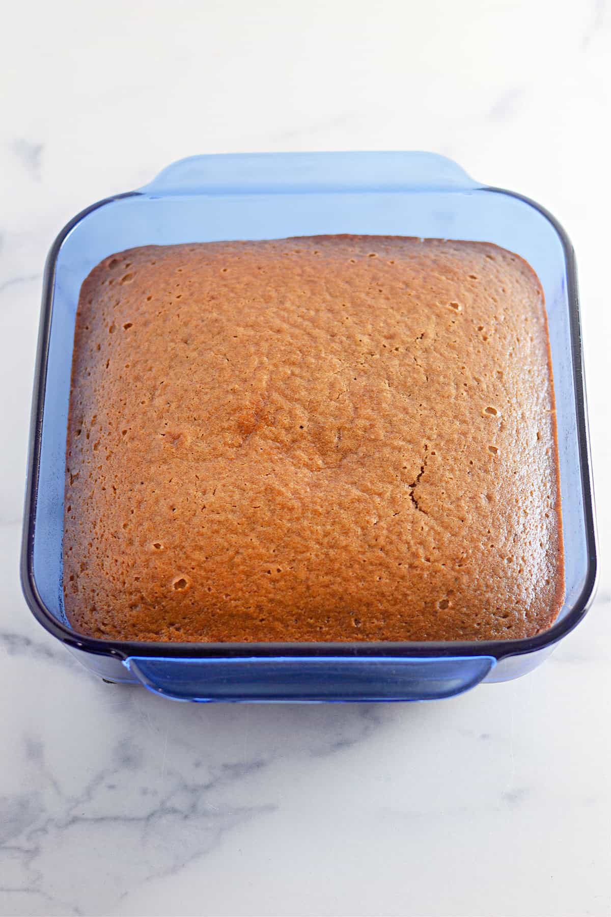 A square, golden-brown Pumpkin Cake is shown in a blue glass baking dish on a white marble surface.