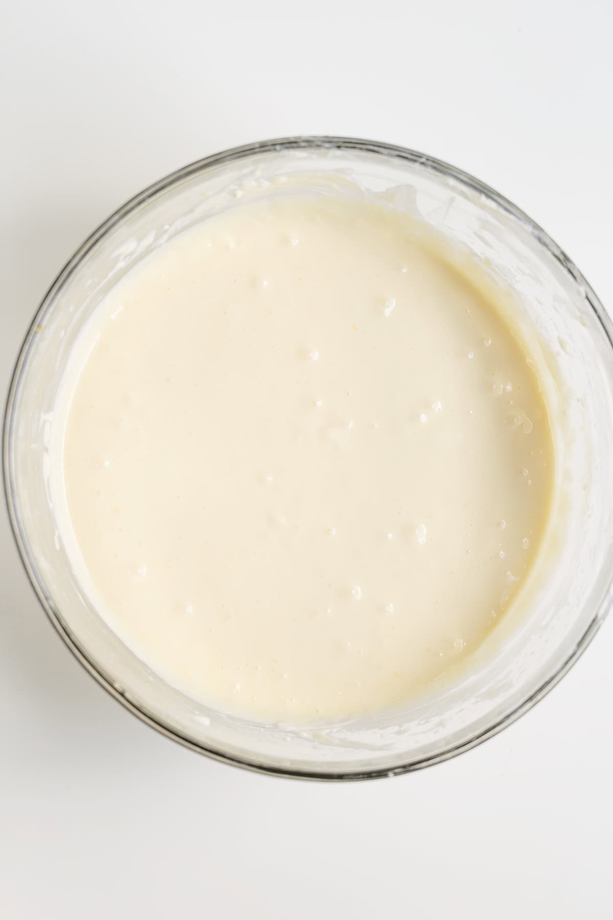 A glass bowl filled with a smooth, pale Raspberry Cheesecake batter sits on a white surface.