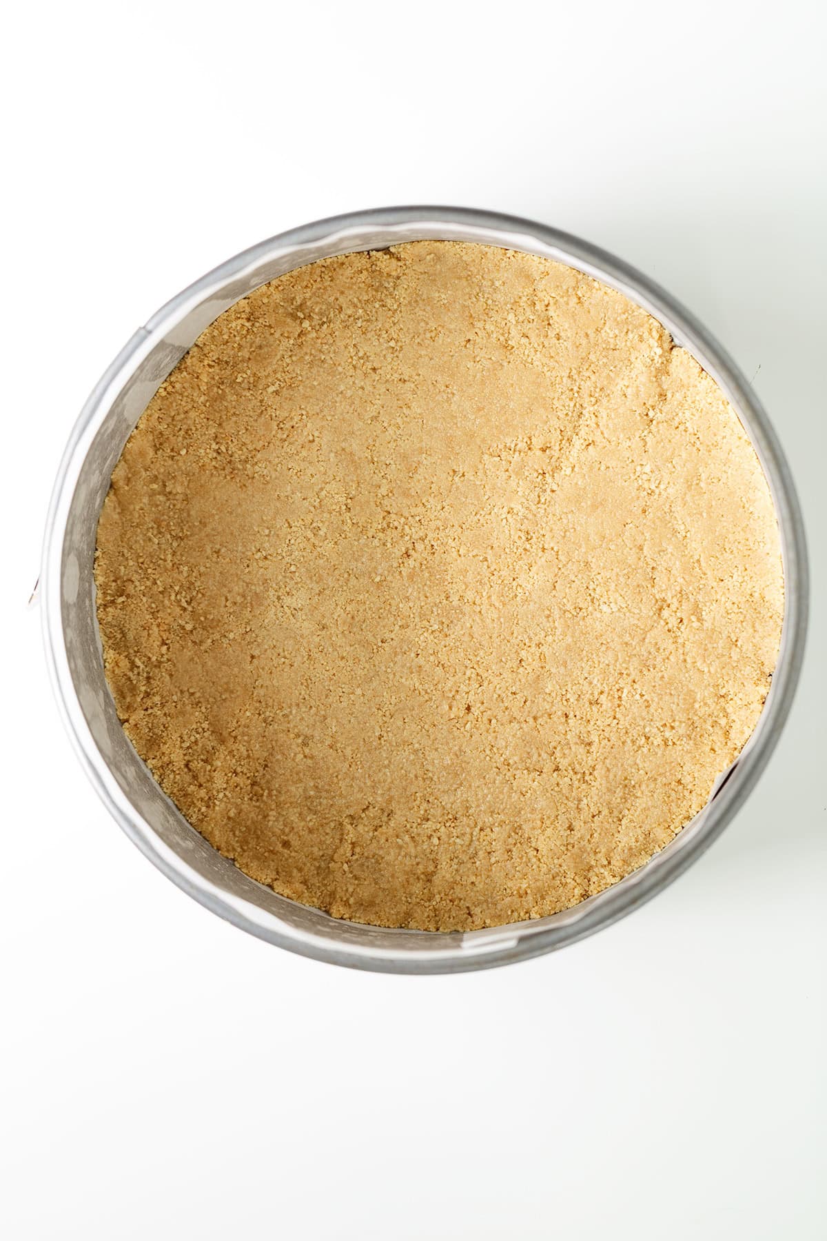 A round baking pan filled with a flat, evenly pressed layer of crushed graham cracker crust on a white surface—perfect as the base for a luscious raspberry cheesecake.