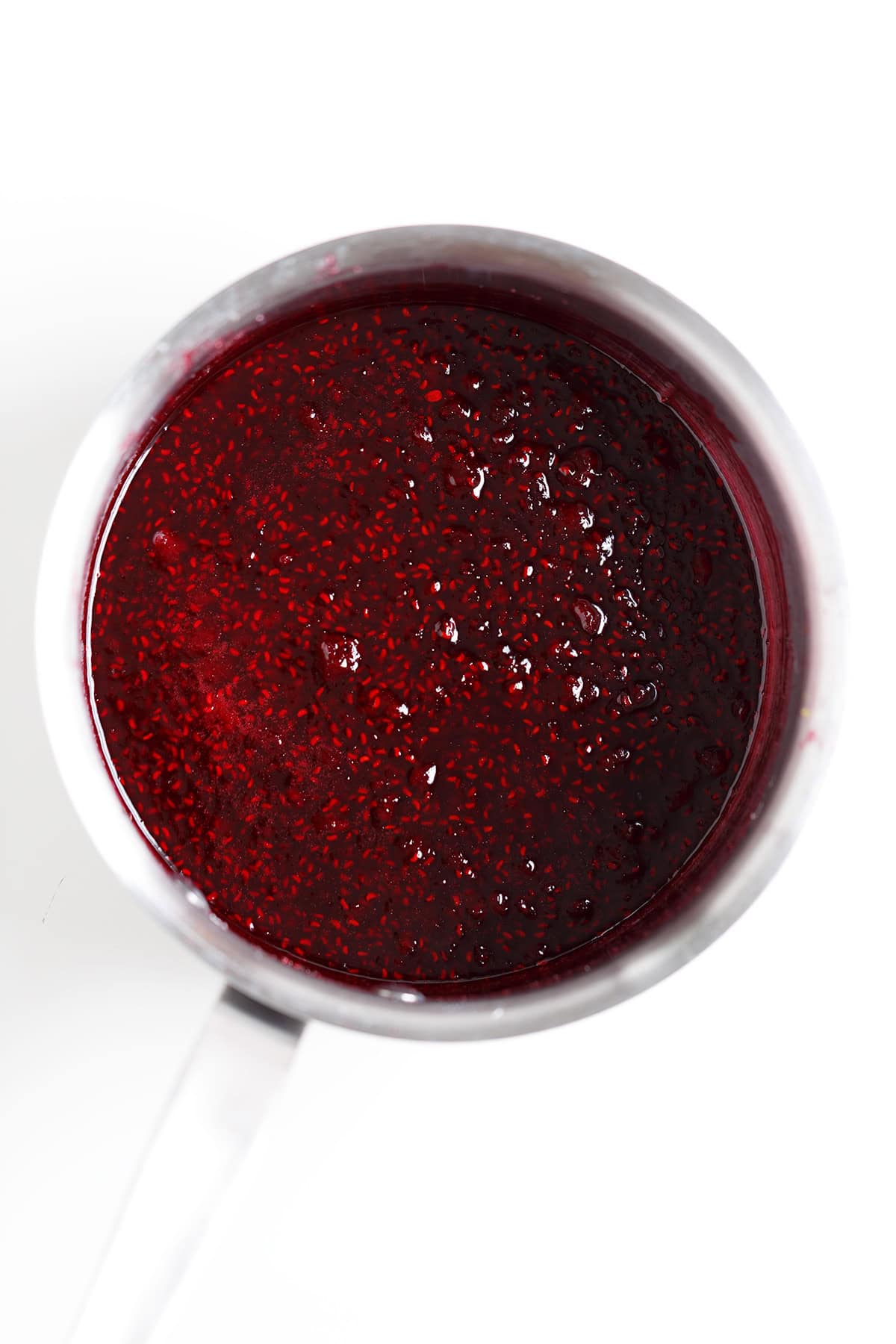 A stainless steel saucepan filled with thick, dark red raspberry sauce or compote—perfect for drizzling over a creamy cheesecake—is viewed from above on a white background.