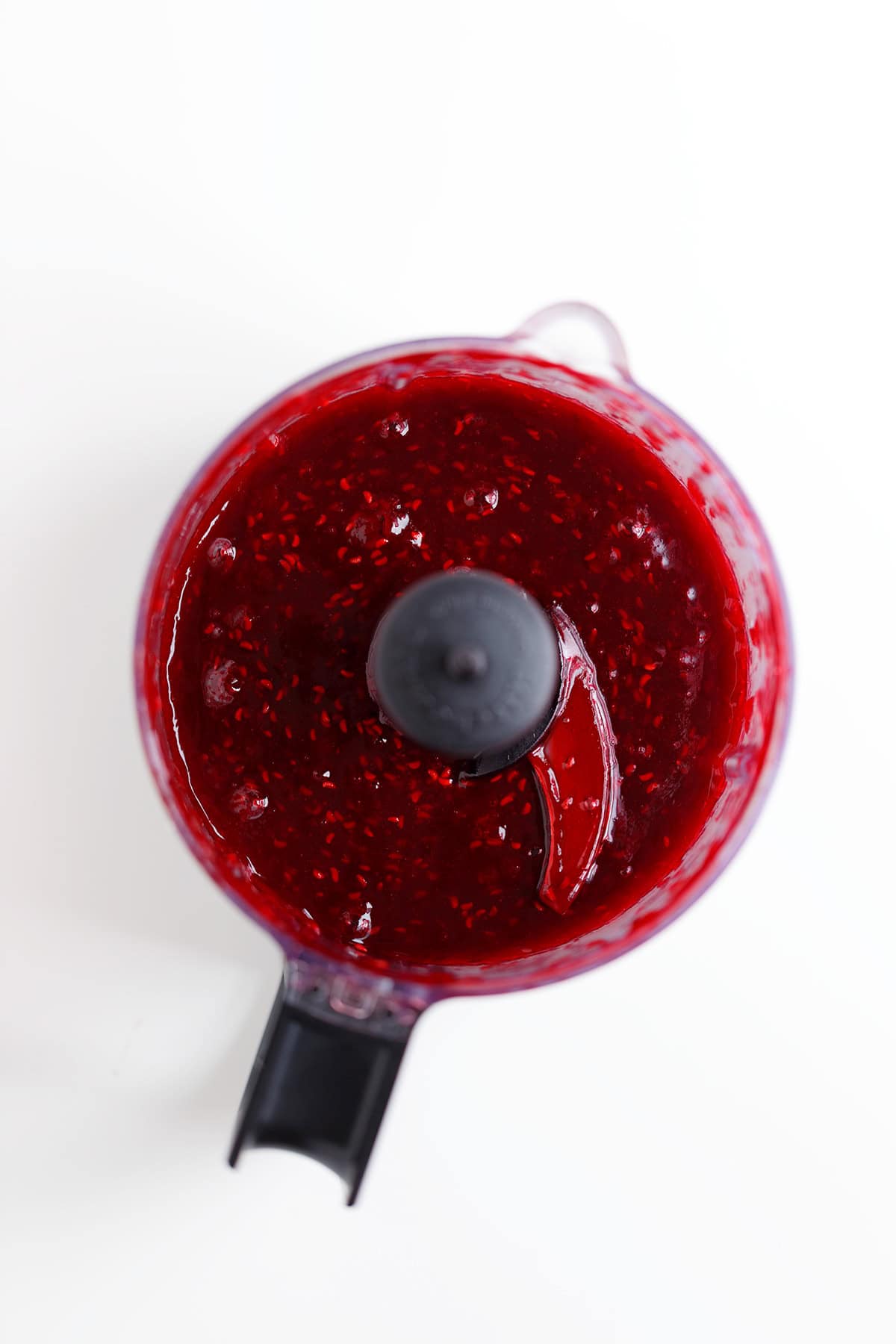 A food processor filled with blended raspberry cheesecake sauce, viewed from above, on a white surface.