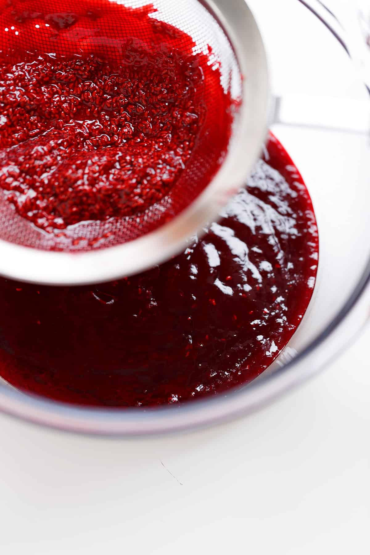 A metal strainer is being used to separate raspberry seeds from a bowl of raspberry sauce or puree, perfect for creating a silky topping for Raspberry Cheesecake.