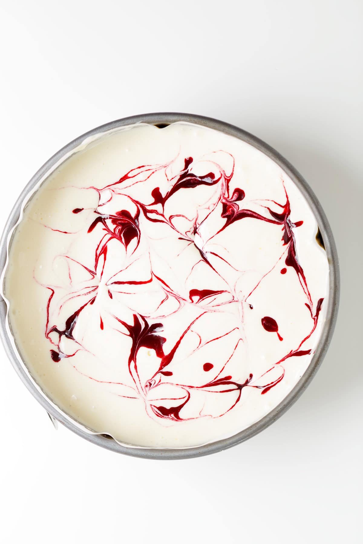 A round pan filled with creamy Raspberry Cheesecake batter, elegantly swirled with vibrant red raspberry sauce on top, viewed from above on a white background.