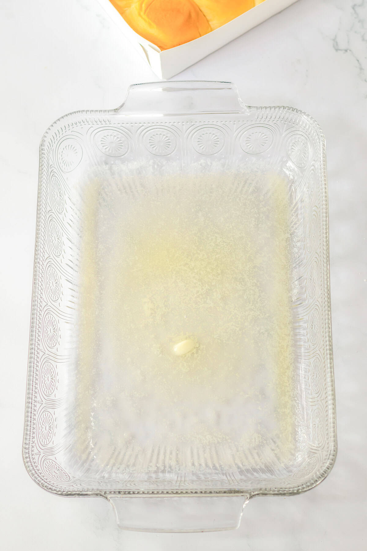 A clear glass baking dish with melted butter in the bottom, sitting on a white surface. In the background, a partially visible box of Hawaiian Rolls with orange contents adds a hint of warmth to the scene.