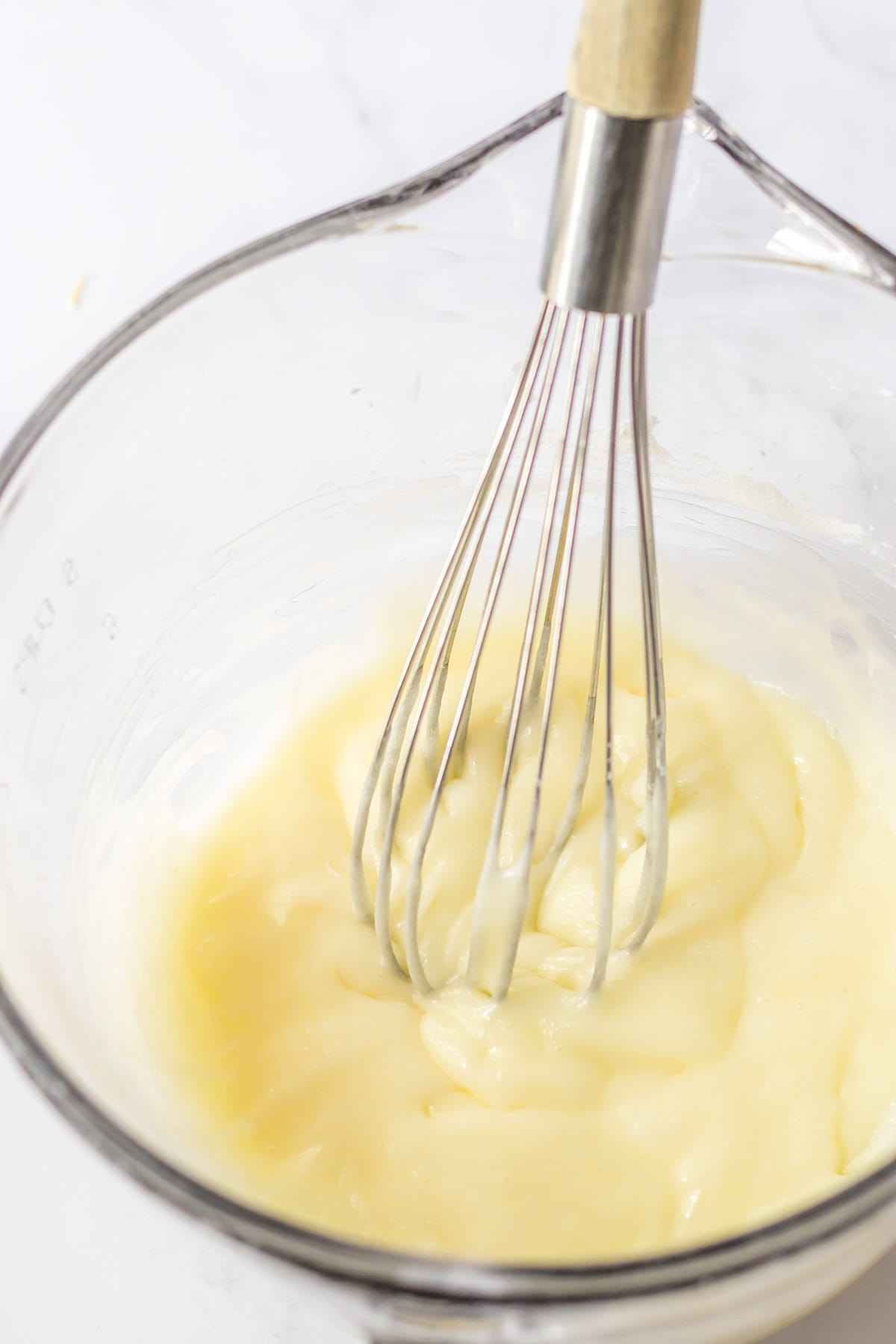 A metal whisk rests in a glass bowl filled with thick, creamy batter for cinnamon rolls on a light surface.
