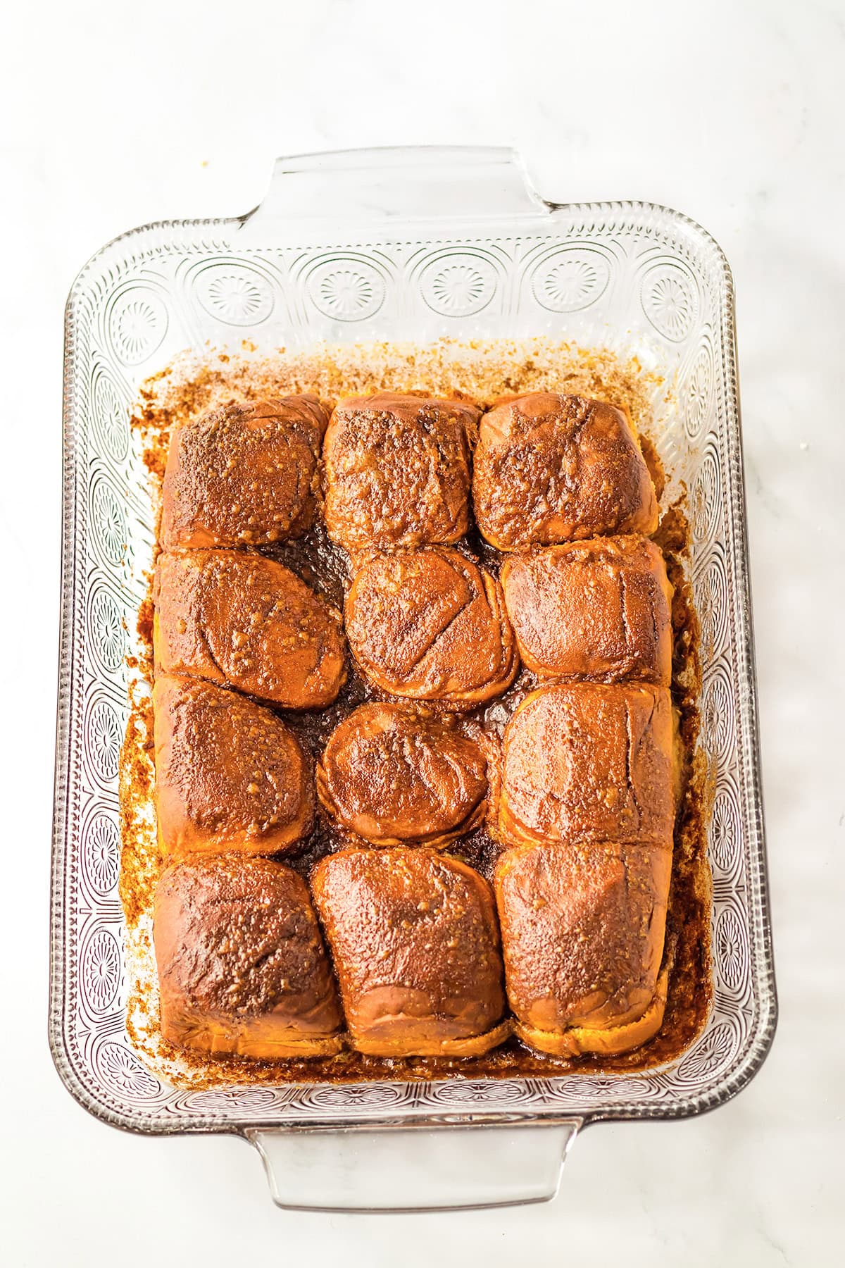 A glass baking dish with twelve golden brown Hawaiian Roll slider buns arranged in three rows, freshly baked and topped with seasoning.