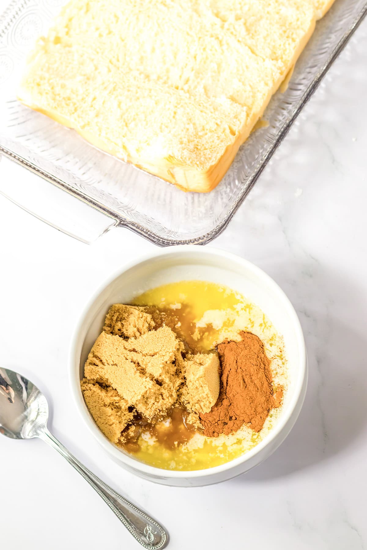 A bowl with brown sugar, melted butter, cinnamon, and vanilla sits next to a freshly baked Hawaiian Roll Cinnamon Roll cake and a spoon on a marble countertop.