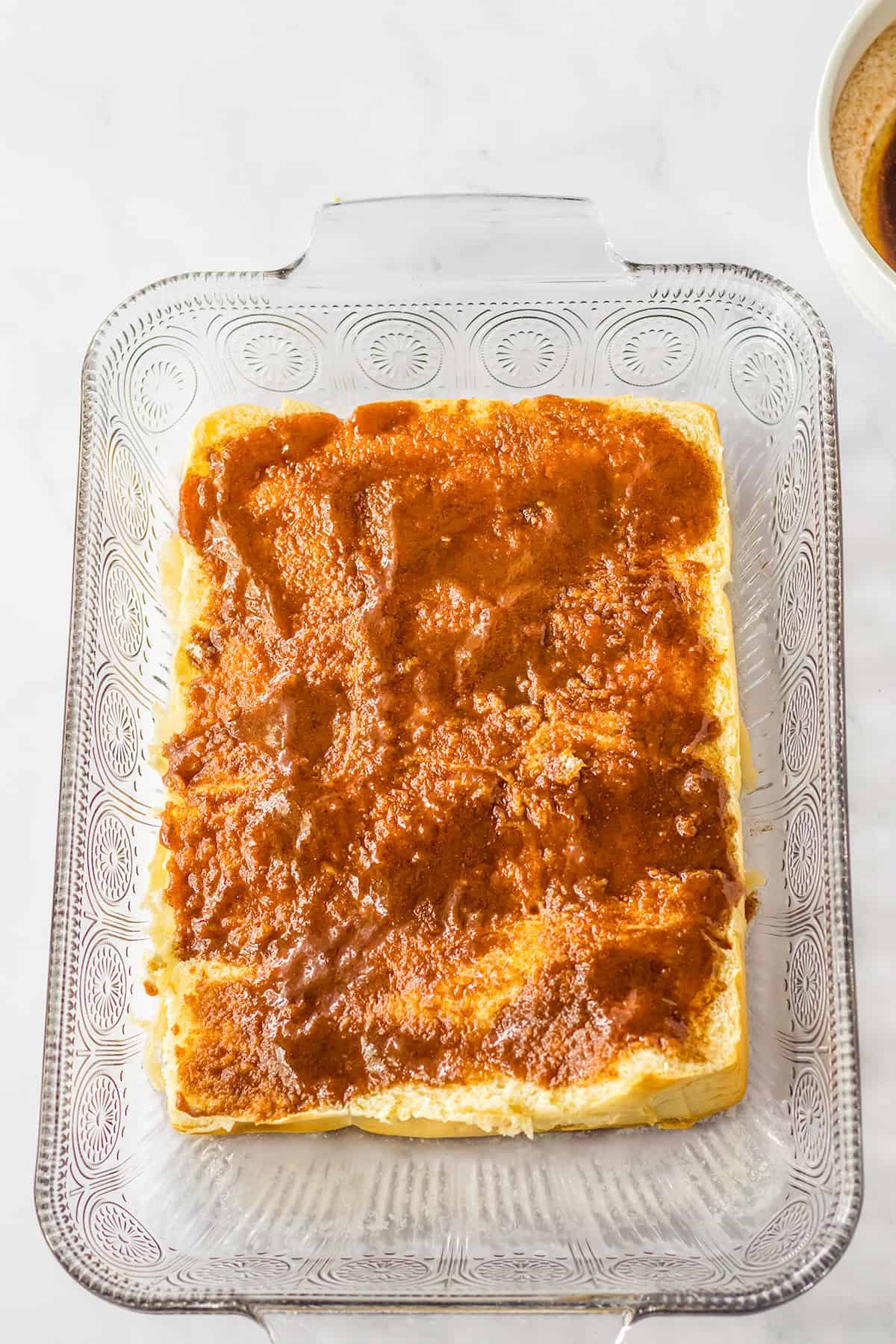 Glass baking dish with a layer of bread, reminiscent of Hawaiian Roll Cinnamon Rolls, topped with a brown, spreadable mixture, set on a white countertop.