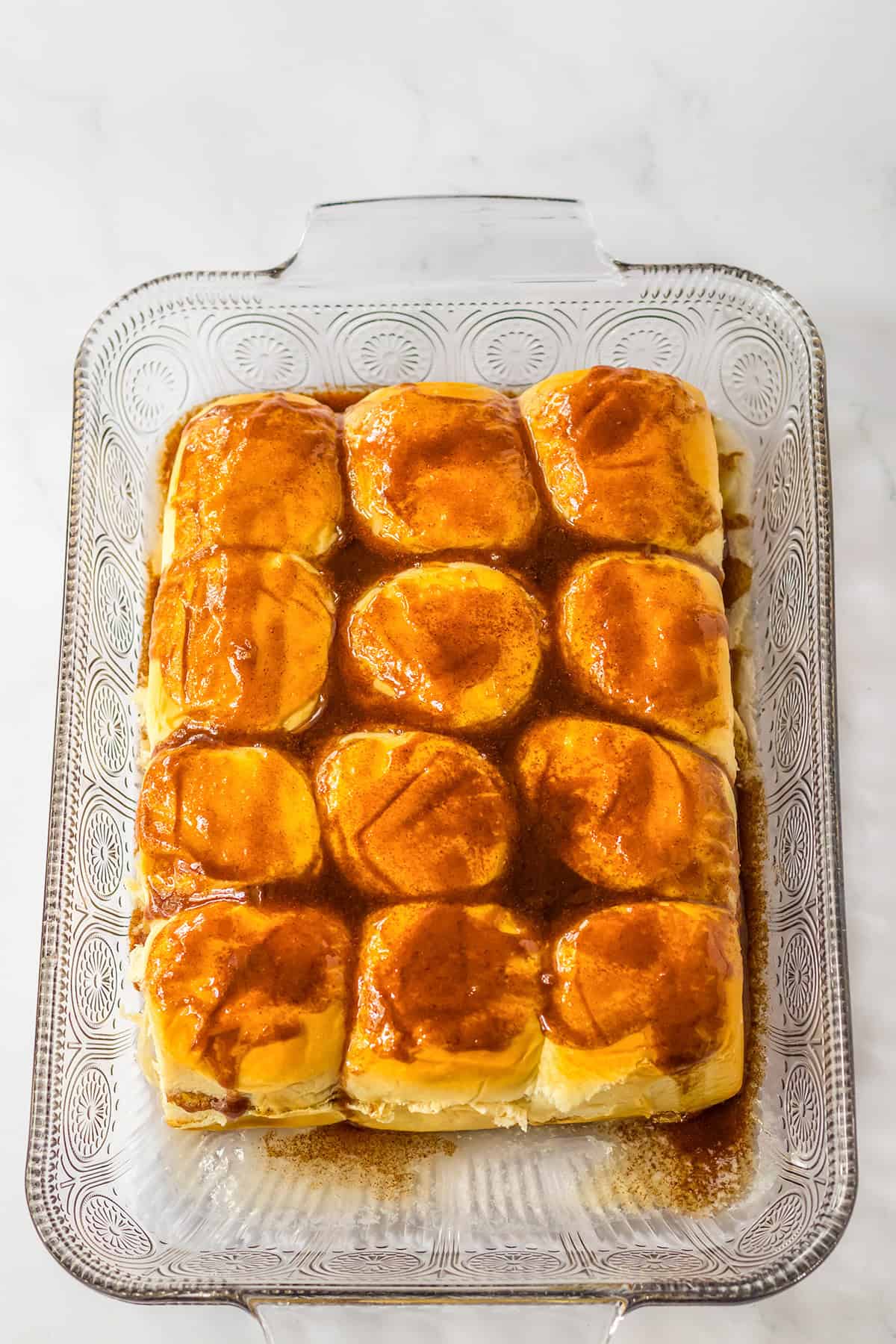 A glass baking dish with twelve golden brown, glazed Hawaiian Roll Cinnamon Rolls arranged in a 3x4 grid on a white surface.