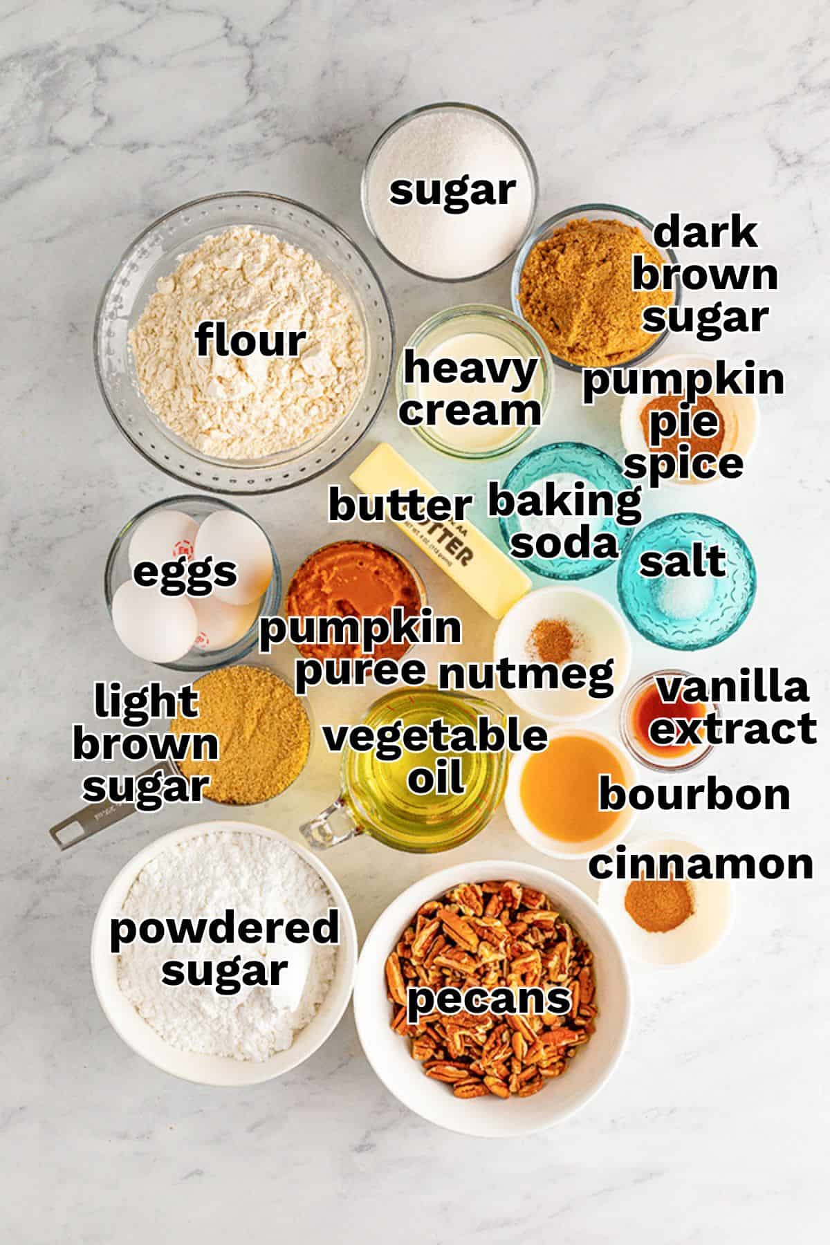 Various baking ingredients, including flour, sugars, eggs, pumpkin puree, heavy cream, spices, oil, vanilla extract, bourbon, powdered sugar, baking soda, salt, and pecans are arranged on a countertop—everything you need to create a delicious Pumpkin Sheet Cake topped with homemade Praline Icing.