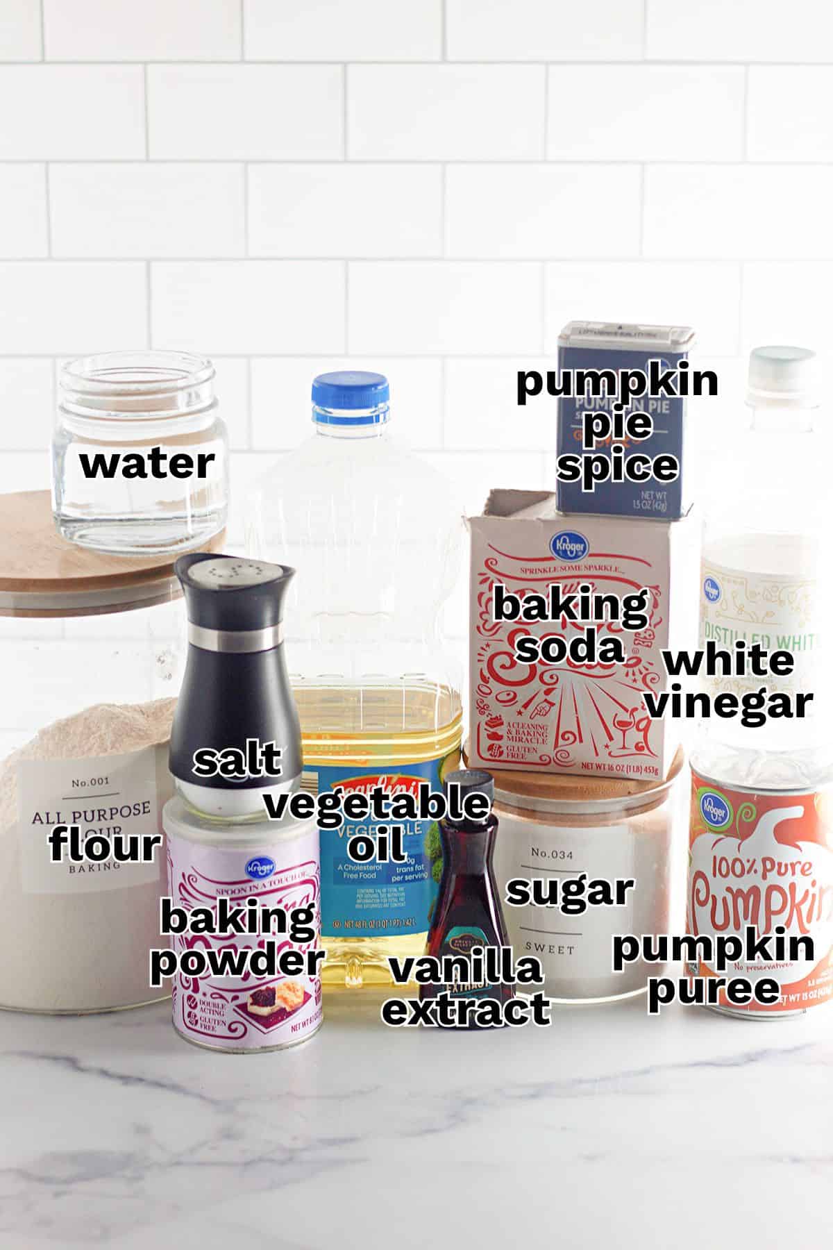 A group of baking ingredients on a counter, including flour, oil, sugar, pumpkin puree, pumpkin pie spice, baking powder, baking soda, salt, vanilla extract, water, and white vinegar—everything you need to create a delicious pumpkin dessert like Pumpkin Wacky Cake.