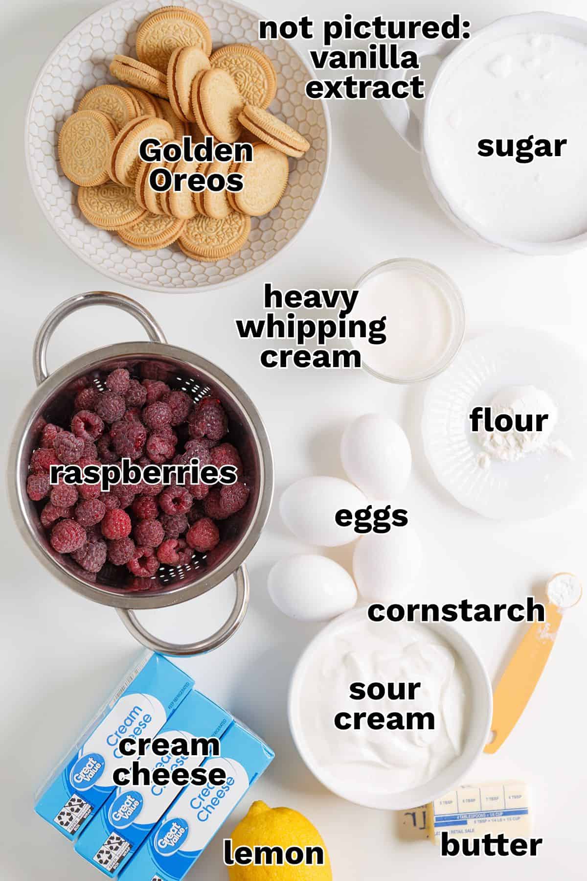 Ingredients for a Raspberry Cheesecake recipe arranged on a white surface, including Golden Oreos, raspberries, cream cheese, lemon, sour cream, eggs, sugar, flour, cornstarch, butter, and cream.