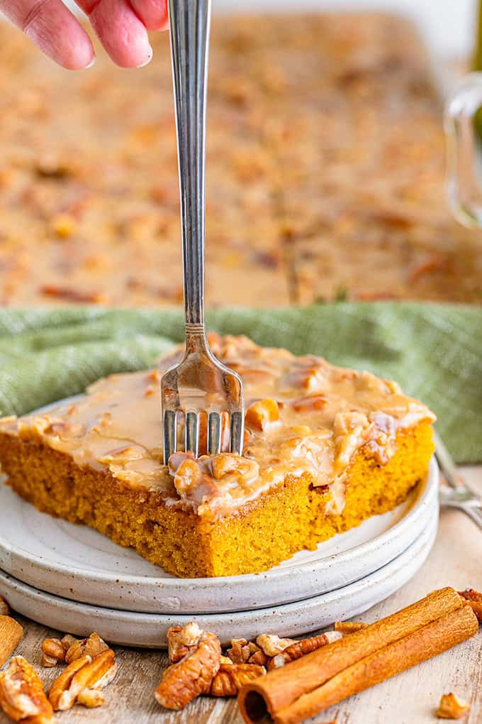 A hand holds a fork about to cut into a slice of pumpkin sheet cake with pecans, topped with luscious praline icing and served on a white plate. Cinnamon sticks and pecans are scattered nearby.