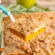 A hand holds a spatula lifting a square piece of Pumpkin Sheet Cake with glazed praline icing and chopped pecans from a larger tray of cake.