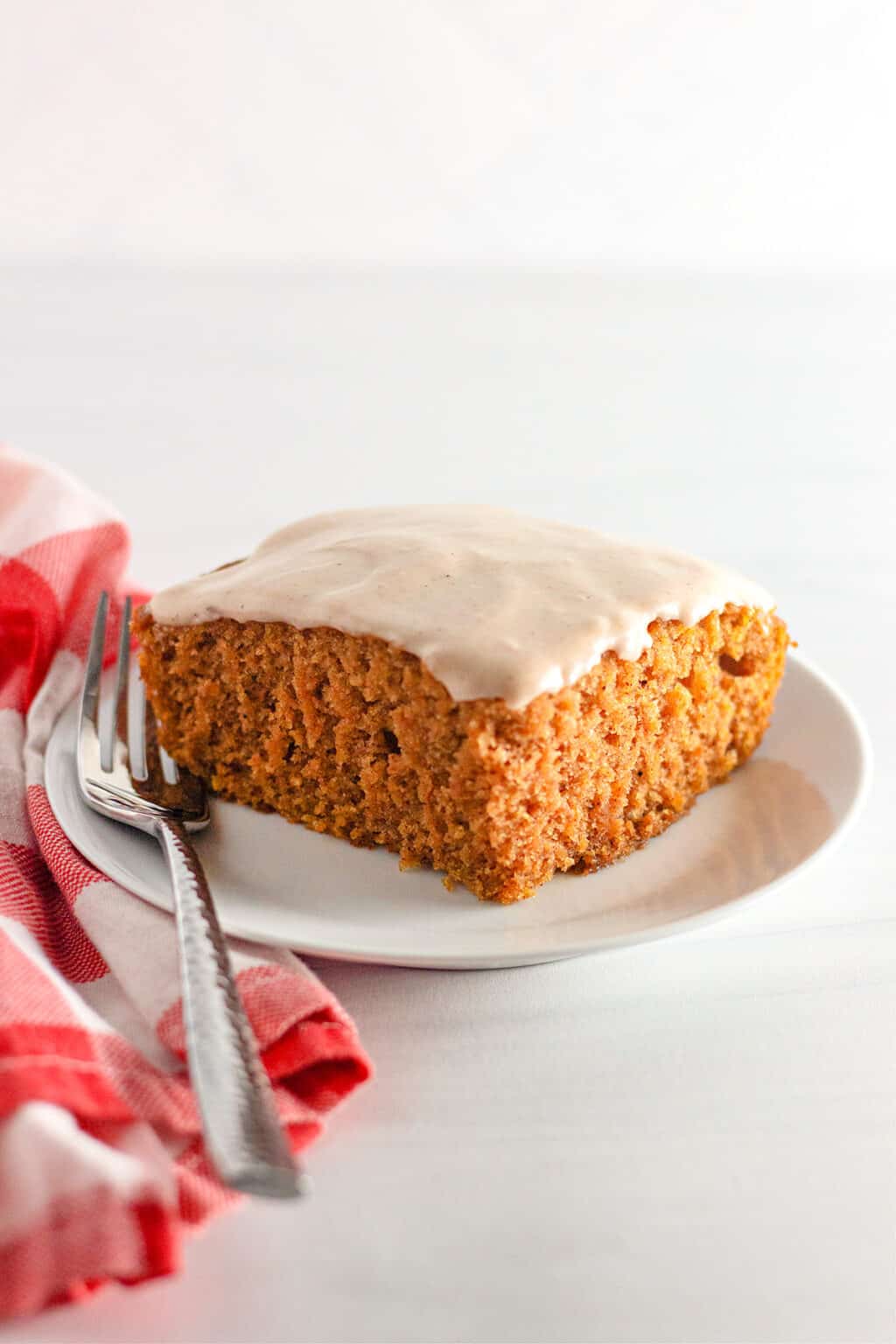 A square piece of frosted Pumpkin Wacky Cake sits on a white plate with a fork, next to a red and white checkered cloth.