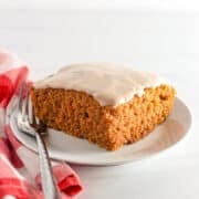 A square piece of frosted Pumpkin Wacky Cake sits on a white plate with a fork, next to a red and white checkered cloth.