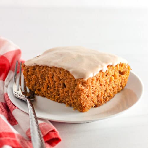 A square piece of frosted Pumpkin Wacky Cake sits on a white plate with a fork, next to a red and white checkered cloth.