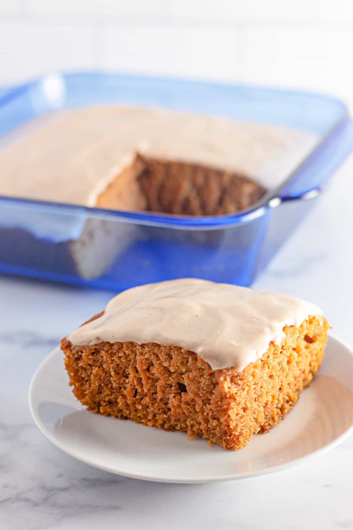 A square piece of frosted Pumpkin Wacky Cake sits on a plate, with the remaining cake in a blue glass baking dish in the background.