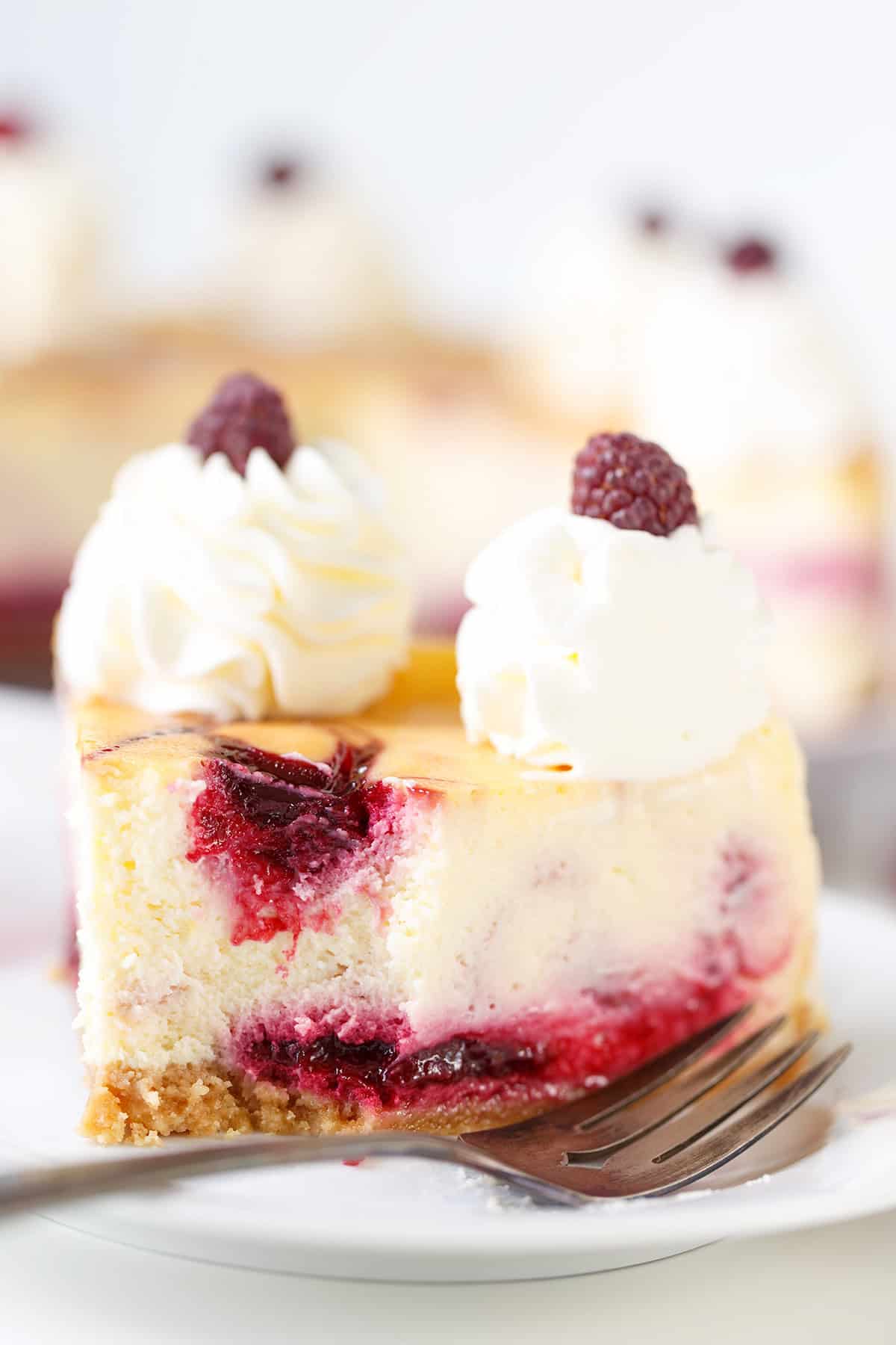 A decadent slice of Raspberry Cheesecake with whipped cream and a fresh raspberry on top sits on a white plate with a fork.