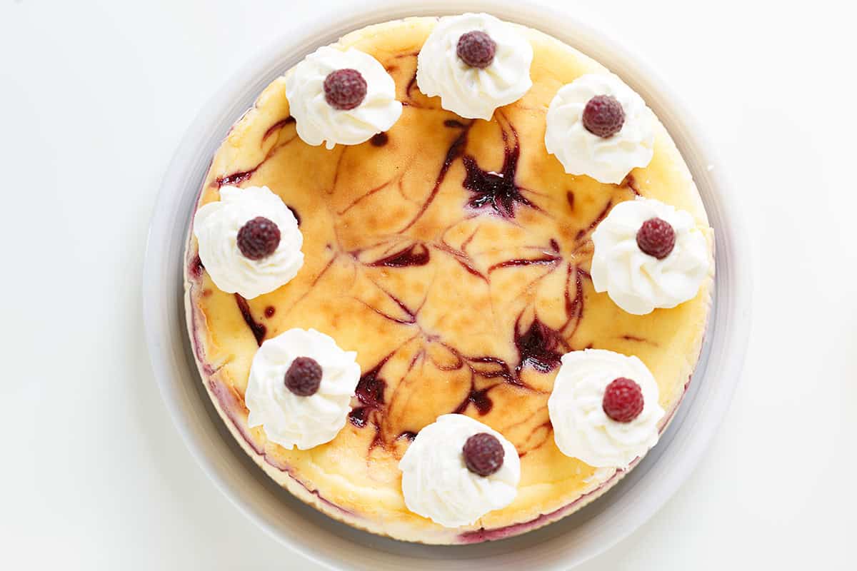 Raspberry cheesecake topped with eight elegant swirls of whipped cream, each garnished with a vibrant raspberry, photographed from above on a white surface—an irresistible raspberry dessert.
