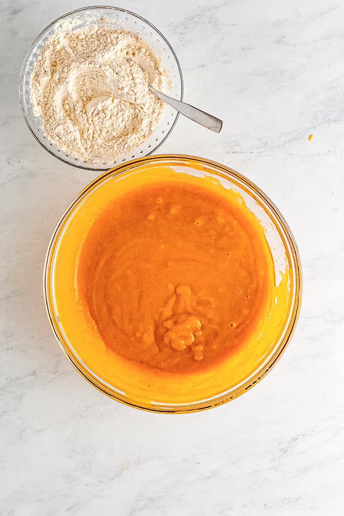 Two glass bowls on a white surface: one with an orange pumpkin mixture for a delicious Pumpkin Sheet Cake, the other with dry flour mixture and a spoon.