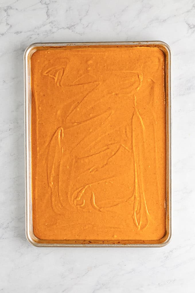 A rectangular sheet pan filled with smooth, unbaked pumpkin sheet cake batter sits on a white marble surface.