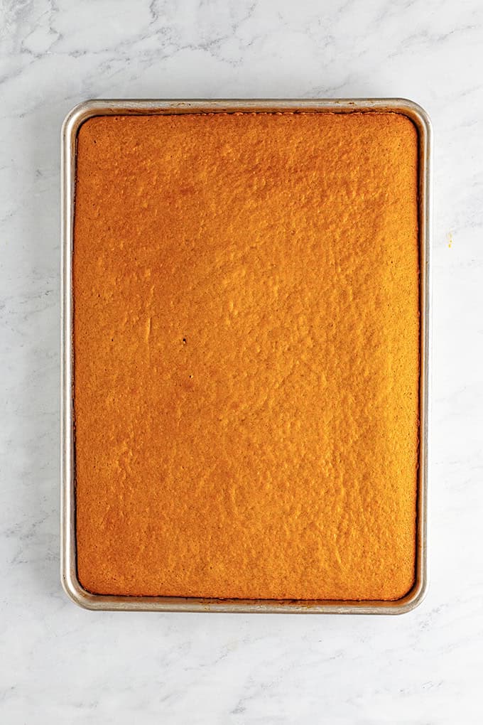 A rectangular Pumpkin Sheet Cake with a golden brown top sits in a metal baking pan on a marble surface.
