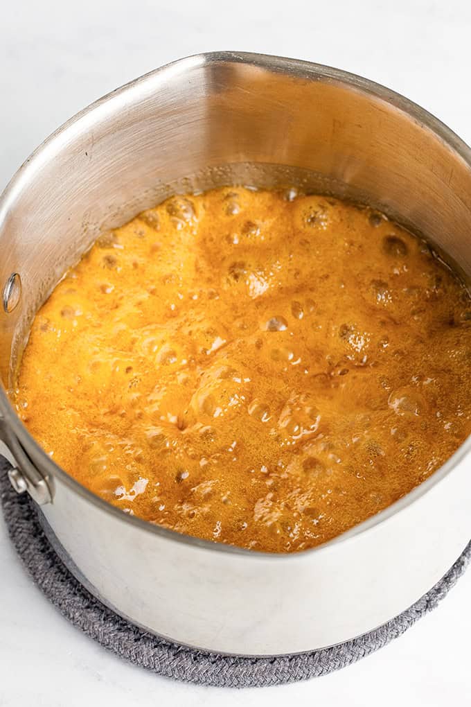 A stainless steel pot filled with bubbling amber-colored liquid, likely praline icing, rests on a gray trivet on a white surface.