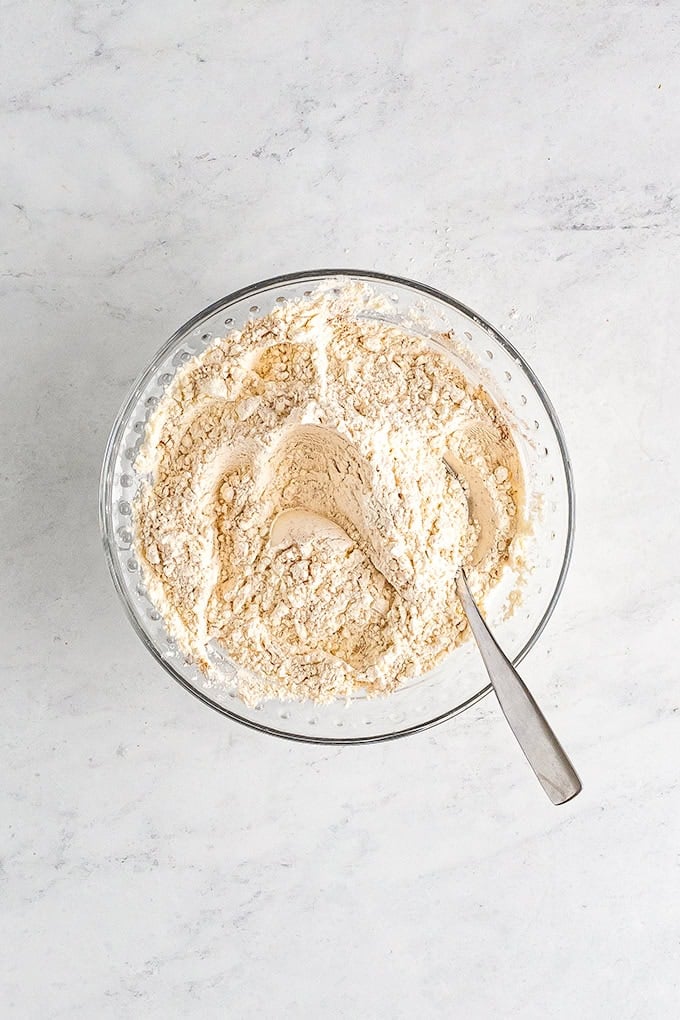A glass bowl filled with flour mixture and a metal spoon, placed on a light-colored marble surface, ready for making a delicious pumpkin sheet cake.