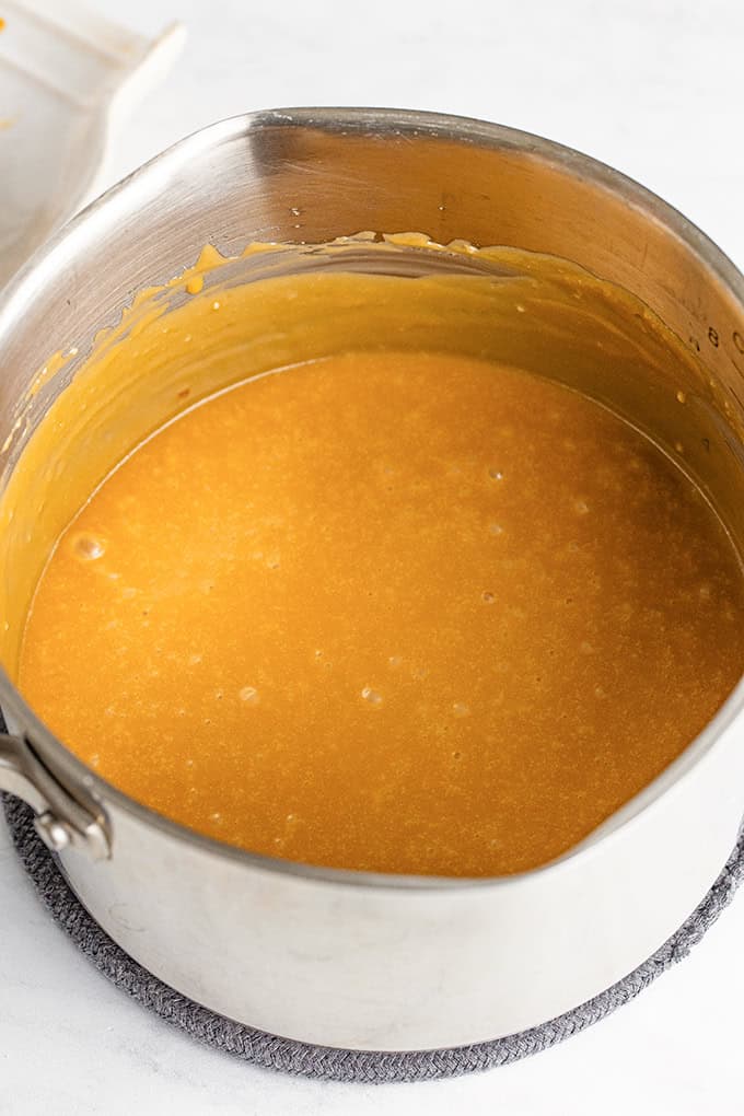 A saucepan filled with a smooth, golden-brown praline icing sits on a gray trivet on a white surface, ready to be drizzled over a freshly baked pumpkin sheet cake.