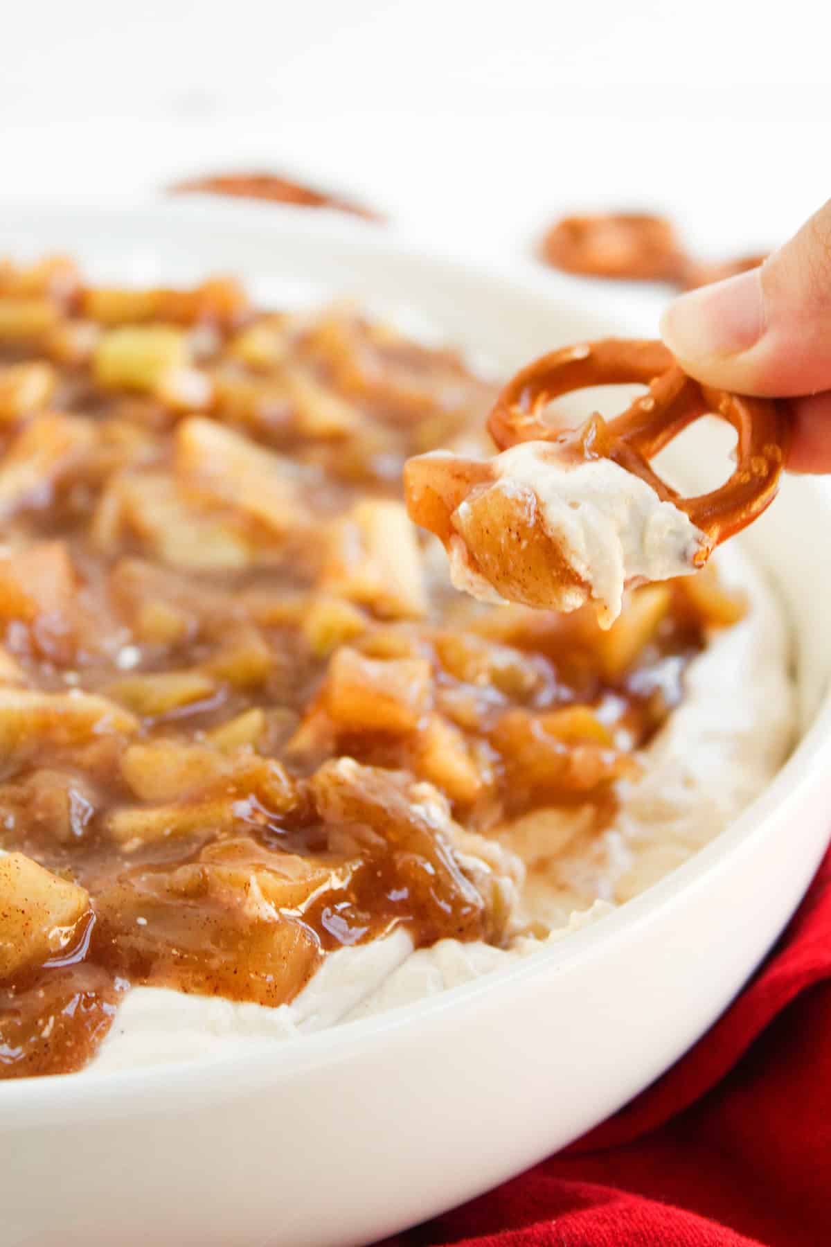 A hand dips a pretzel into a luscious apple pie dip—a creamy dessert dip topped with spiced apple chunks in a white dish.