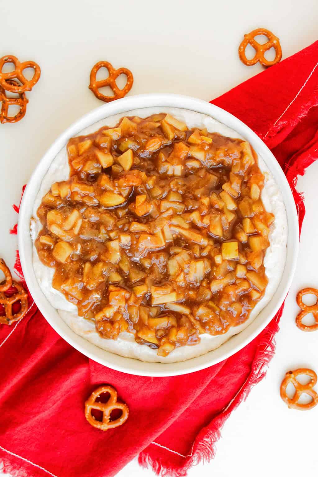 A bowl of apple pie dip with a rich, cream cheese and brown, chunky topping sits on a red napkin, surrounded by scattered pretzels on a white surface.