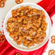 A bowl of apple pie dip with a rich, cream cheese and brown, chunky topping sits on a red napkin, surrounded by scattered pretzels on a white surface.