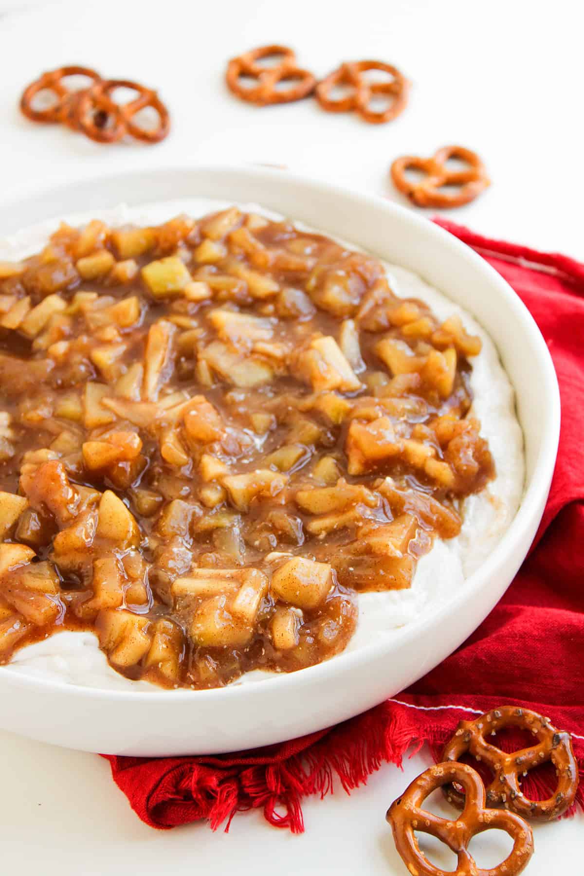 A white dish filled with creamy cream cheese dip topped with a chunky apple cinnamon mixture&mdash;reminiscent of a delicious Apple Pie Dip&mdash;is surrounded by pretzels on a red napkin, making for a delightful apple dessert.