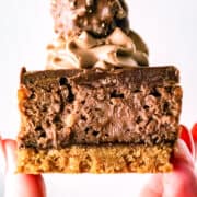 A hand holds a decadent Ferrero Rocher Cheesecake bar with a cookie base, chocolate cheesecake layer, chocolate cream topping, and a round Ferrero Rocher candy on top.