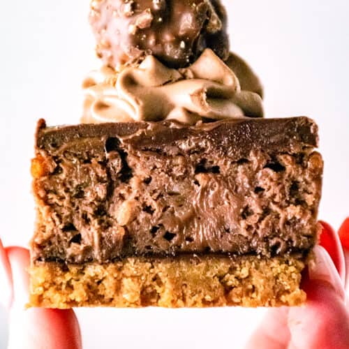 A hand holds a decadent Ferrero Rocher Cheesecake bar with a cookie base, chocolate cheesecake layer, chocolate cream topping, and a round Ferrero Rocher candy on top.