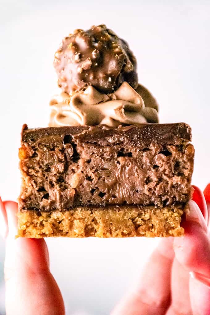 A hand holds a decadent Ferrero Rocher Cheesecake bar with a cookie base, chocolate cheesecake layer, chocolate cream topping, and a round Ferrero Rocher candy on top.