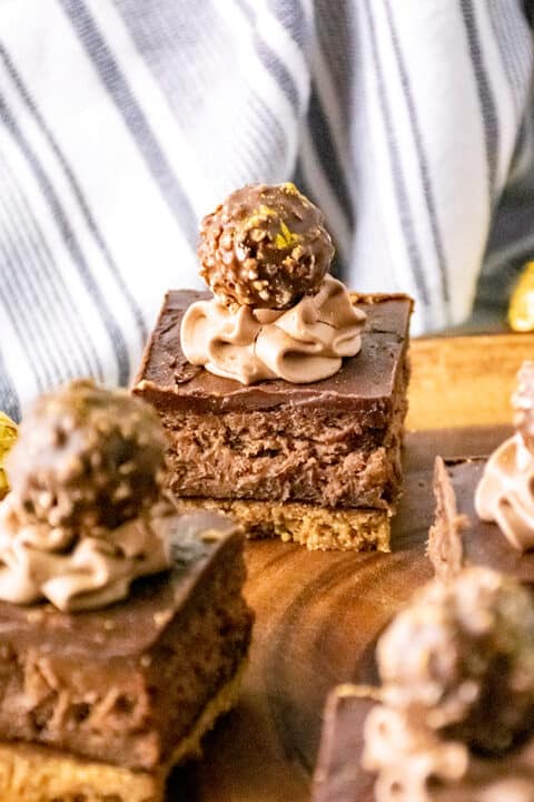 A chocolate dessert bar with a creamy layer, reminiscent of decadent cheesecake bars, is topped with rich chocolate frosting and crowned with a Ferrero Rocher candy, sitting elegantly on a wooden board.