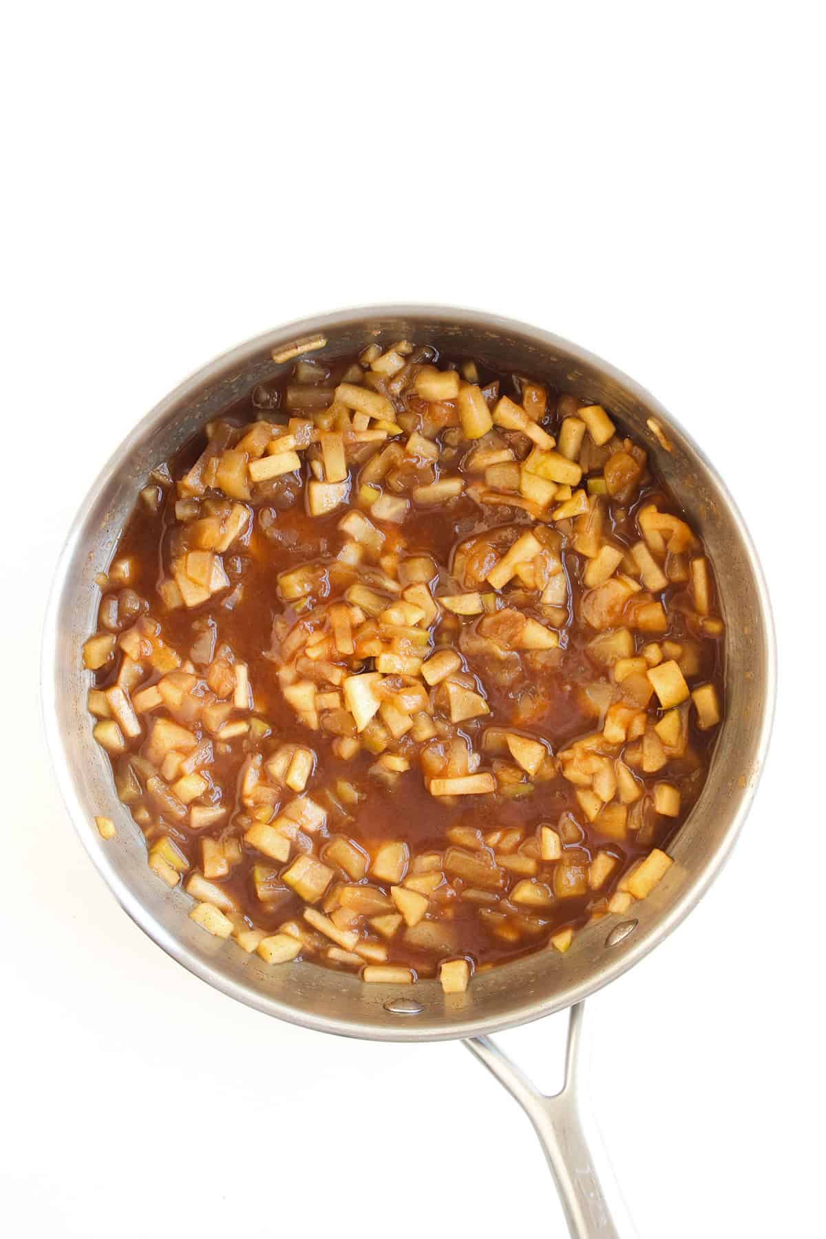 A stainless steel saucepan filled with diced apples cooked in a brown, syrupy sauce—perfect for making a warm Apple Pie Dip—is viewed from above against a white background.