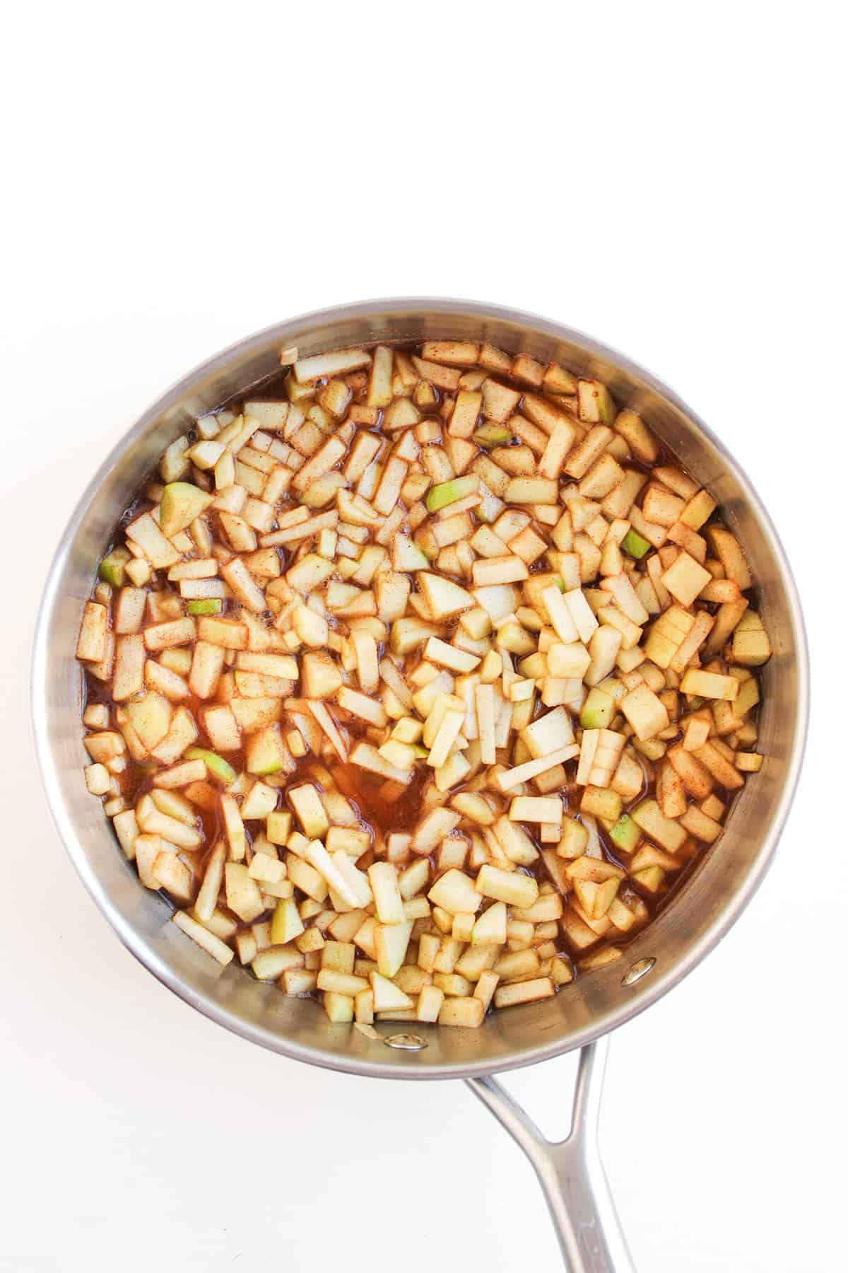 A stainless steel pan filled with diced apples coated in a brown liquid, possibly a mixture of cinnamon and sugar, sits on a white background—perfect for creating an Apple Pie Dip to serve with Cream Cheese.