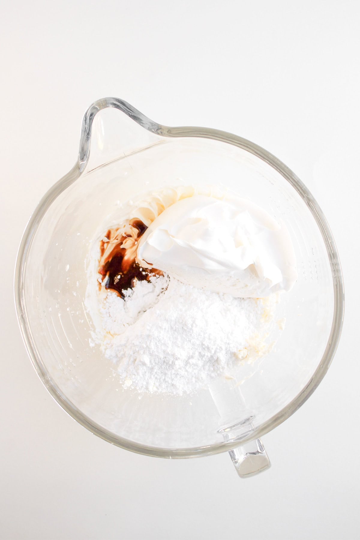 A glass mixing bowl containing whipped topping, powdered sugar, cream cheese, and vanilla extract on a white surface—perfect for making a luscious cream cheese dip to pair with apple pie dip.