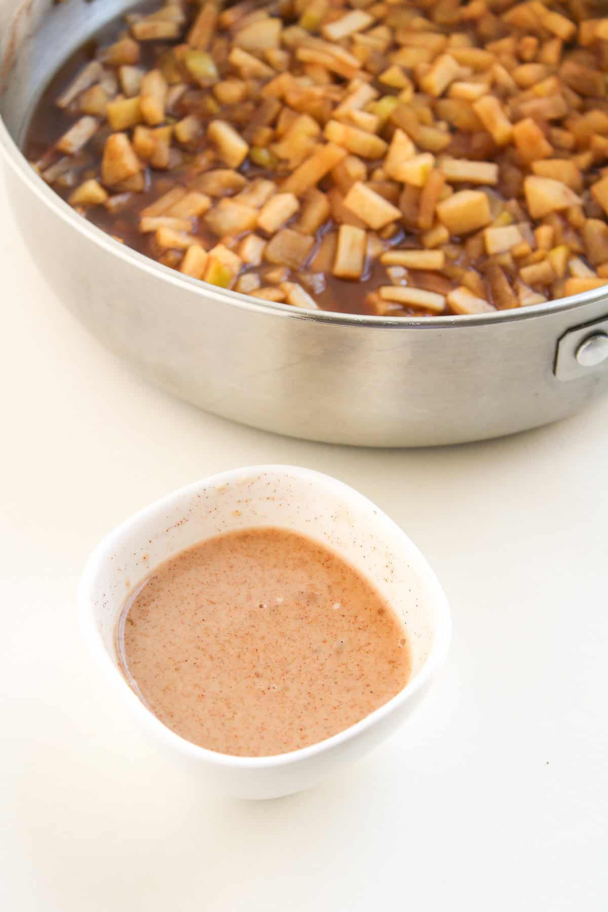 A small bowl of cinnamon apple pie dip sits in front of a pan filled with diced apples cooking in liquid, both placed on a white surface.