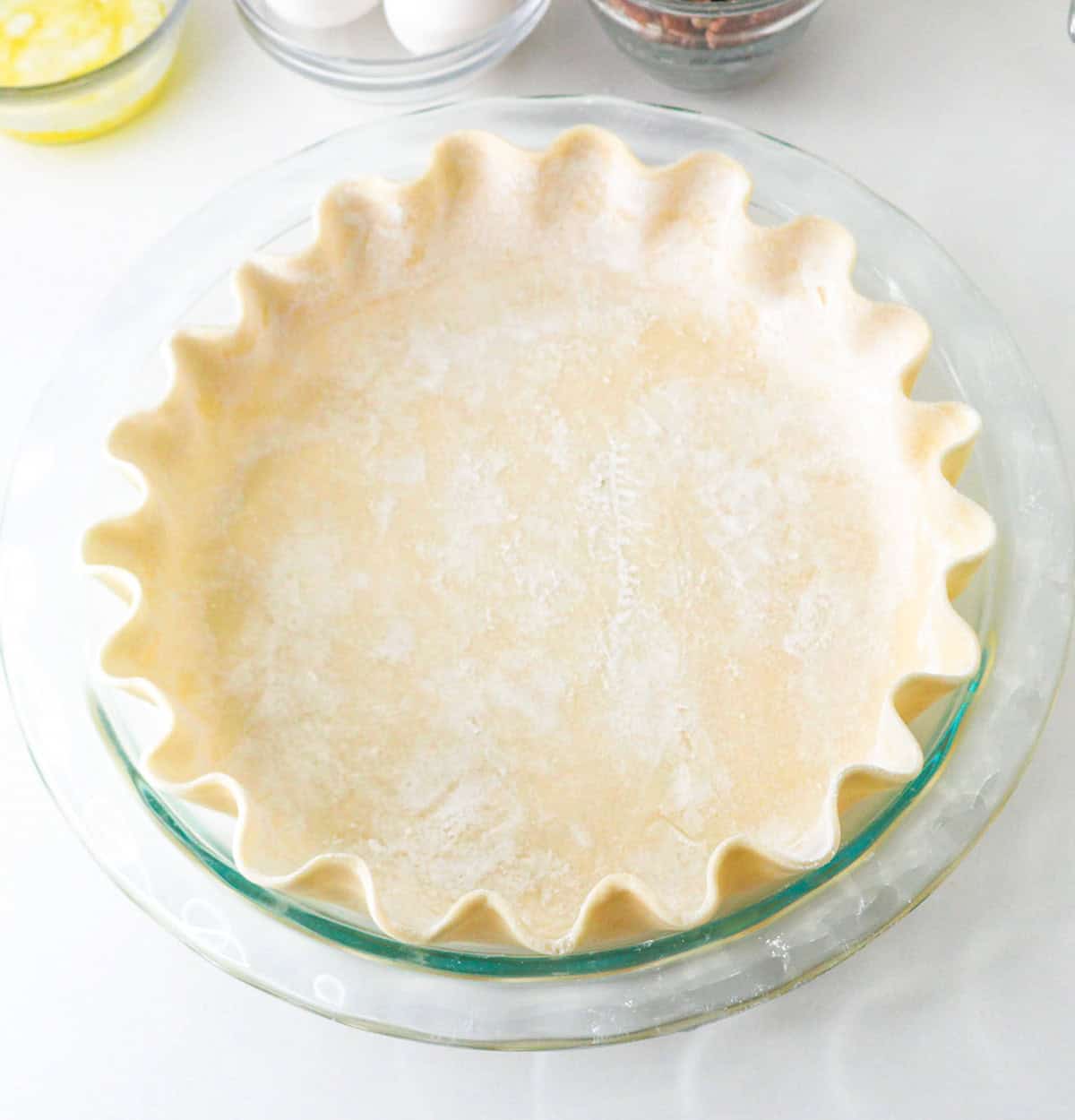 Unbaked pie crust with crimped edges in a glass pie dish, perfect for preparing a delicious Pumpkin Pecan Pie, placed on a white surface with baking ingredients nearby.